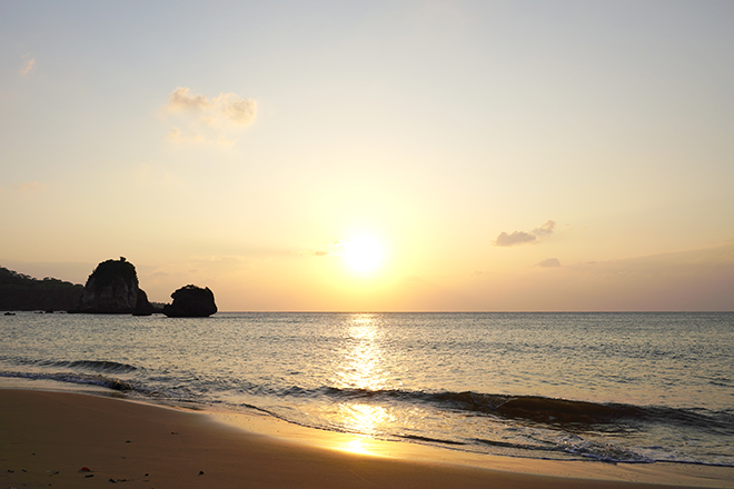 浜辺で沈みゆく太陽と、オレンジがかった穏やかな海面が太陽光を反射しています